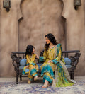 A woman and a young girl wearing matching traditional South Asian outfits pose together on an ornate bench. The outfits are primarily yellow and turquoise blue with intricate embroidery and a sheer patterned dupatta.