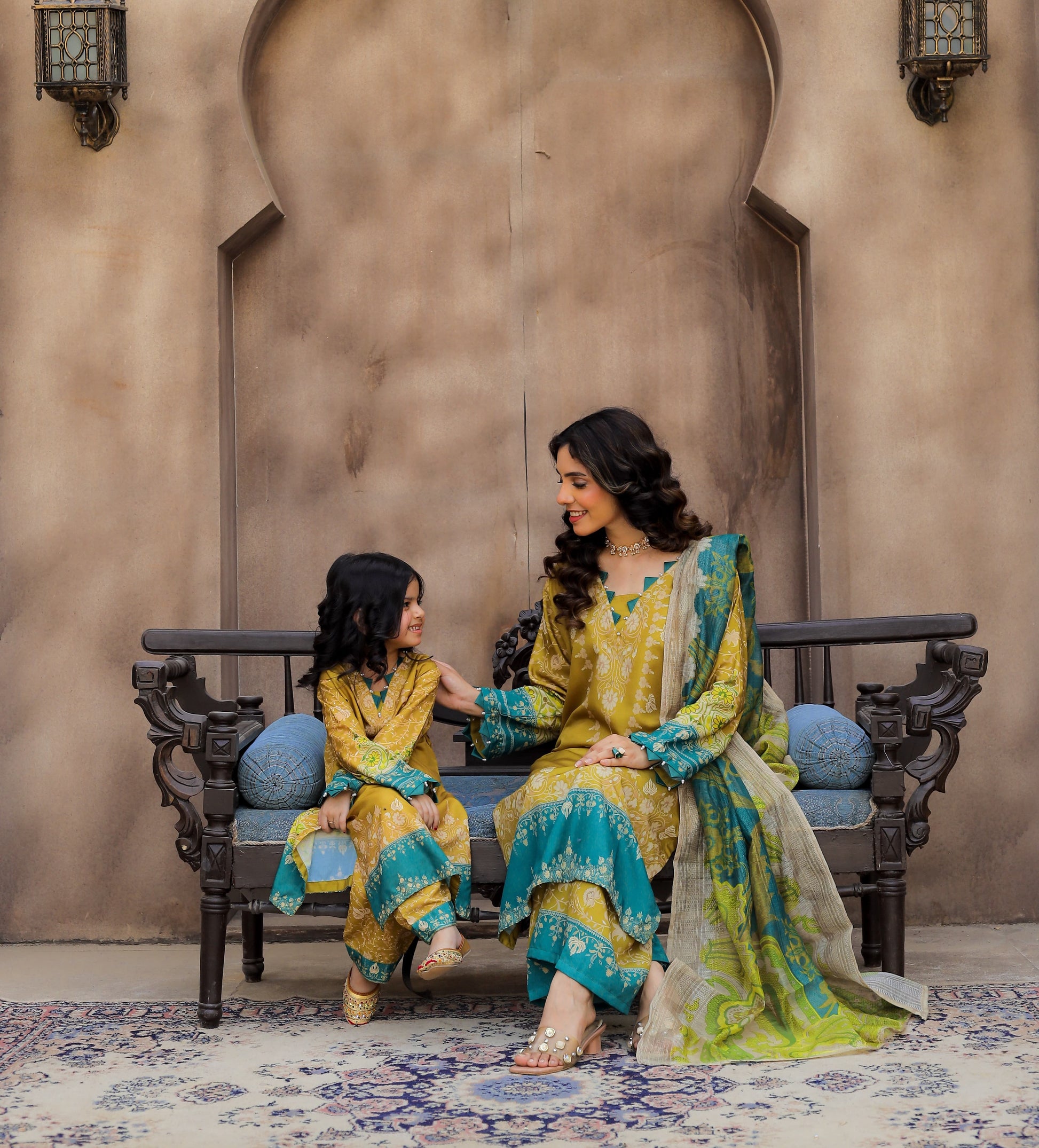 A woman and a young girl wearing matching traditional South Asian outfits pose together on an ornate bench. The outfits are primarily yellow and turquoise blue with intricate embroidery and a sheer patterned dupatta.